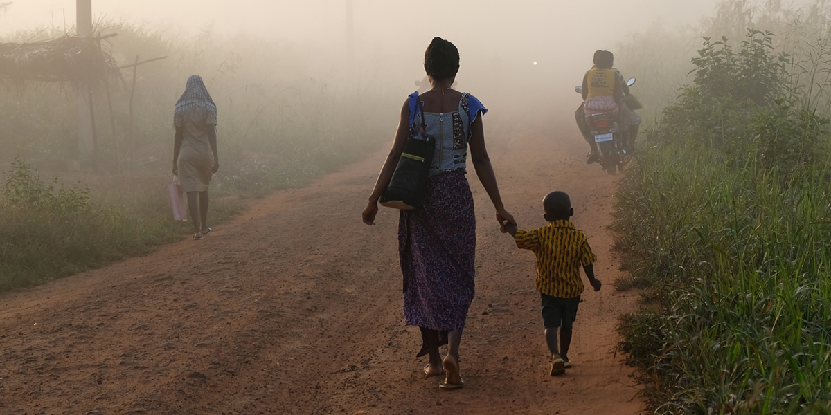 Eine Mutter geht mit ihrem Kind an der Hand eine sandige Straße in Benin, Dogbo entlang